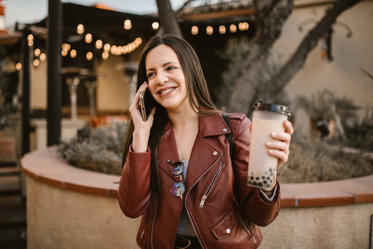 Smiling woman in leather jacket holding bubble tea and talking on the phone outdoors.